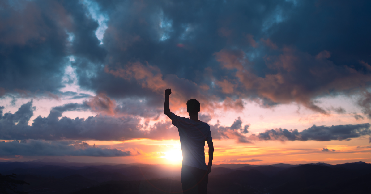 A man triumphantly faces a sunset with his fist raised up in the air.