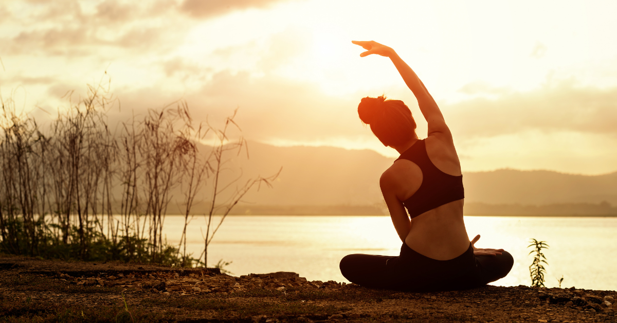 A woman does yoga in the sunlight facing a body of water.