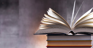 An open book with pages fanned out rests on top of a stack of other books. The background is a textured wall.