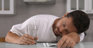 A man passed out over a table with a glass and pills.