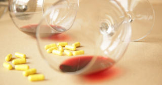 Two wine glasses sit on a surface, one tipped over with red wine spilled. Yellow pills are scattered around the glasses.