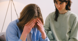 Woman comforts a man who is in distress with his head in his hands.
