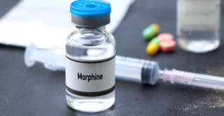 A vial of morphine with a blue cap sits next to a syringe on a dark surface. There are also some colorful pills and a piece of paper visible in the background.