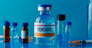 Fentanyl vial in focus, surrounded by syringes and other vials on a reflective blue surface. The word "FENTANYL" is clearly labeled on the vial.