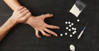 One hand grabs another that is reaching for white pills, a baggy of white powder, and a spoon.