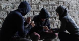 Three people in black hoodies hunch down and pass around unknown substances.